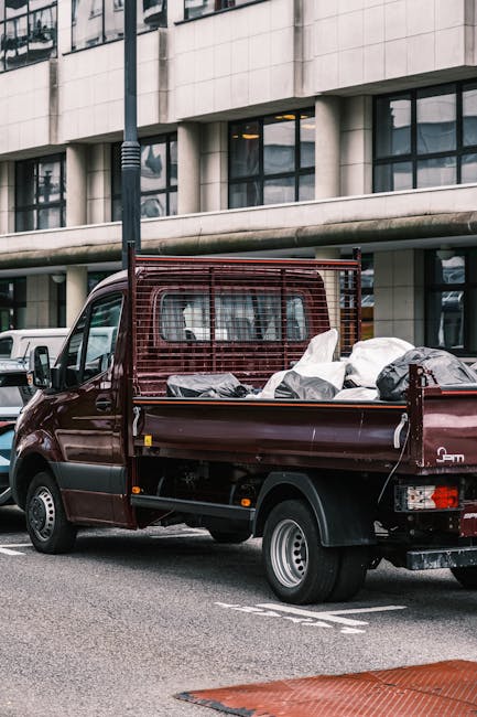 A small, vintage blue pickup truck parked parallel to a curb in an outdoor setting during daytime, loaded with a large amount of mixed waste materials and household rubbish. The truck's body shows sig
