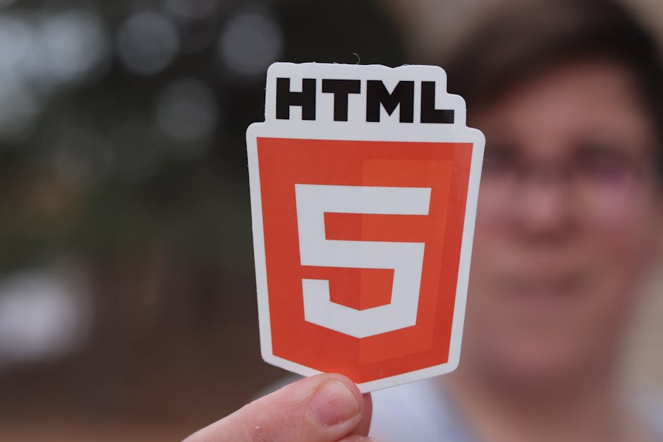 A person holding a sticker featuring the HTML5 logo, which is a red-orange shield with a white numeral '5' and the text 'HTML' above it. The individual is outdoors, with a blurred background of trees